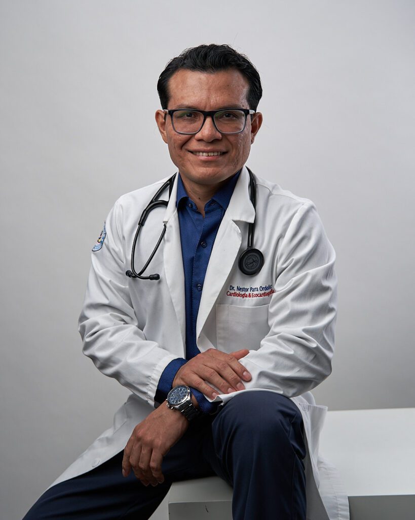 Médico sentado con bata y estetoscopio sentado derecho erguido y la mano derecha apoyada sobre su muñeca izquierda con seguridad.