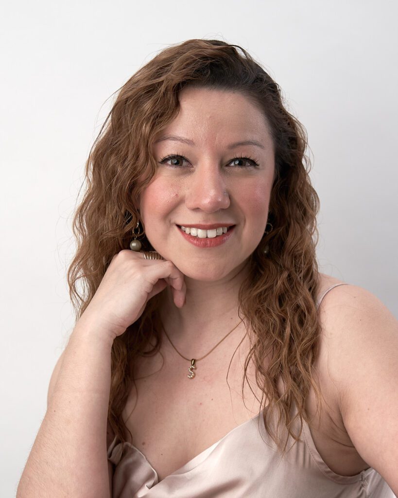 Mujer de cabello castaño ondulado mirando con la cara sonriente mirando a la cámara y apoyada sobre su mano derecha. Ejemplo de Foto Perfil LinkedIn para no Fotogénicos